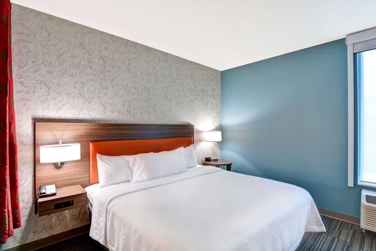 A hotel room with a large bed, two bedside tables with lamps, a nightstand with a phone, and a window with curtains.