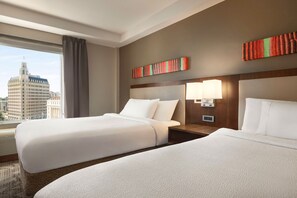 A hotel room with two beds, a large window with a city view, a wall-mounted lamp, and a modern headboard.