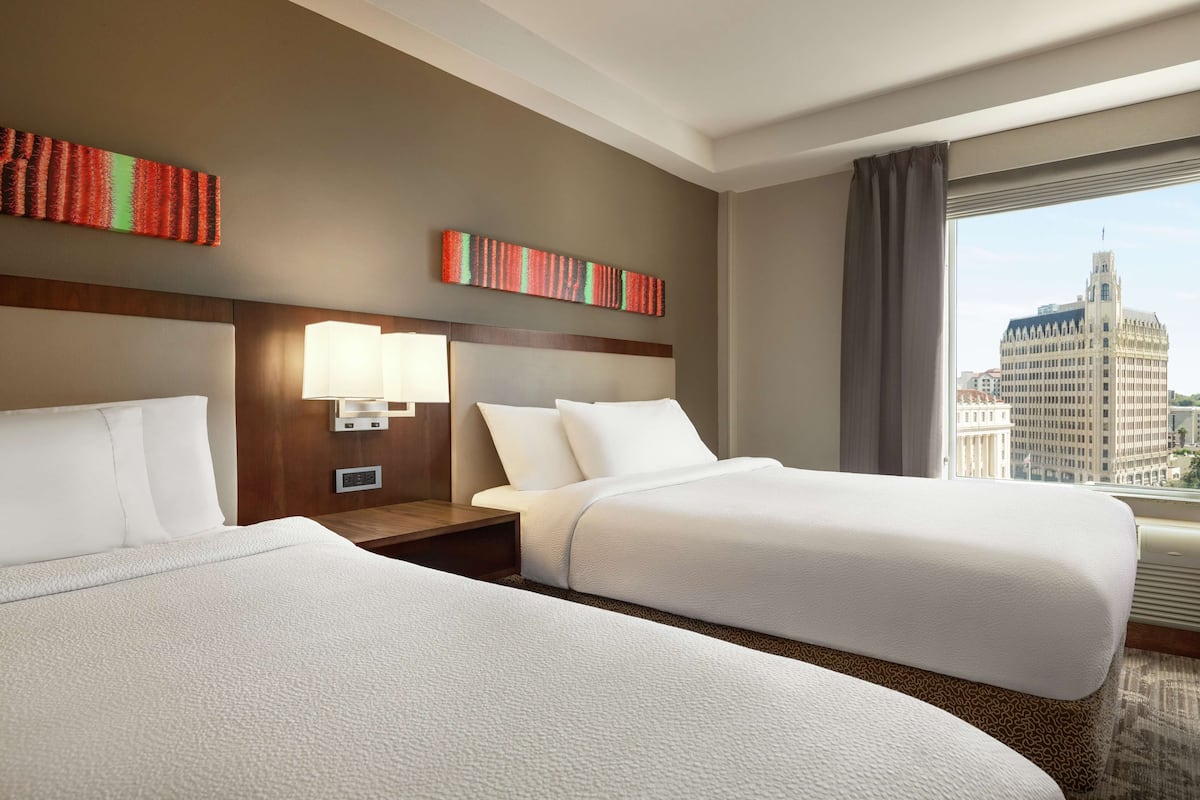 Habitación de hotel con dos camas, una mesita de noche con lámpara, un reloj en la pared y vistas a la ciudad.