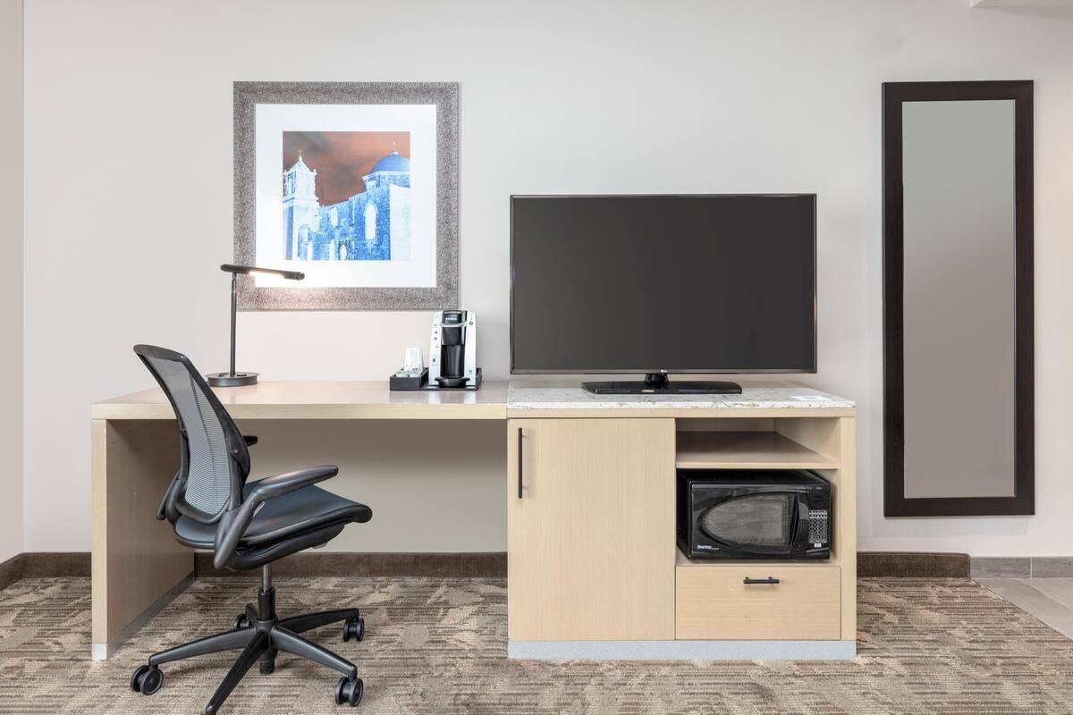 Habitación de hotel con un escritorio de madera, una silla de oficina negra, un televisor de pantalla plana, una cafetera, un microondas y un cuadro en la pared.