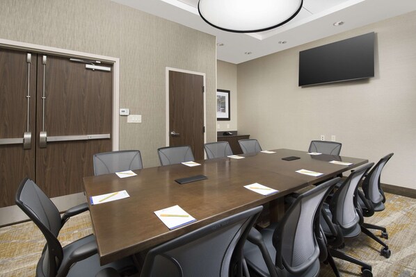 A conference room with a long table, multiple chairs, a flat-screen TV, and wooden doors.