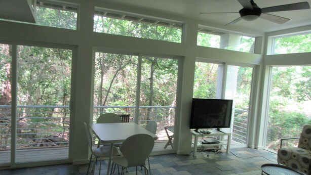Une pièce spacieuse dotée de grandes fenêtres, d’un ventilateur de plafond, d’un téléviseur à écran plat et d’un coin repas avec des chaises et une table.