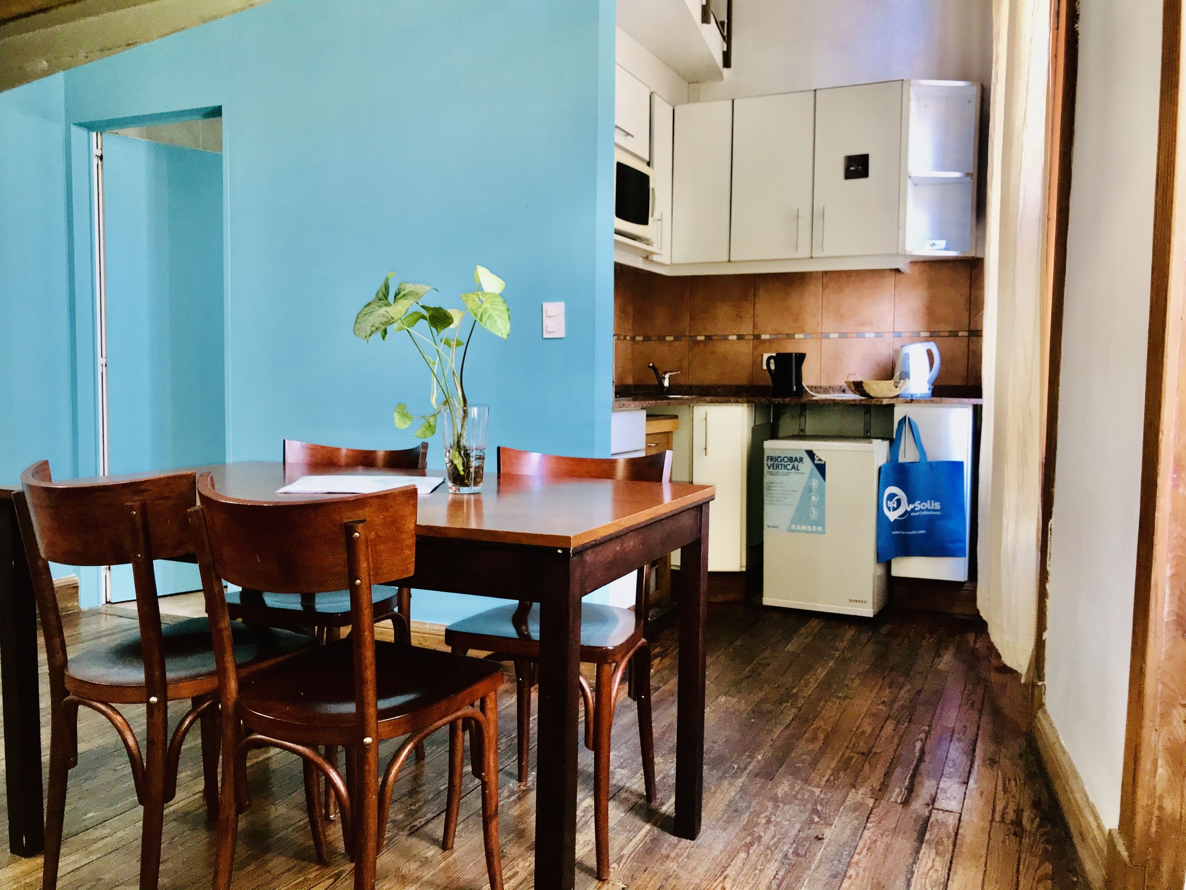A dining area with a wooden table and chairs, a vase with a plant, and a kitchen with white cabinets and a refrigerator.
