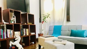 Ein Wohnzimmer mit grauem Sofa, weißem Hocker, Bücherregal und Fernseher.