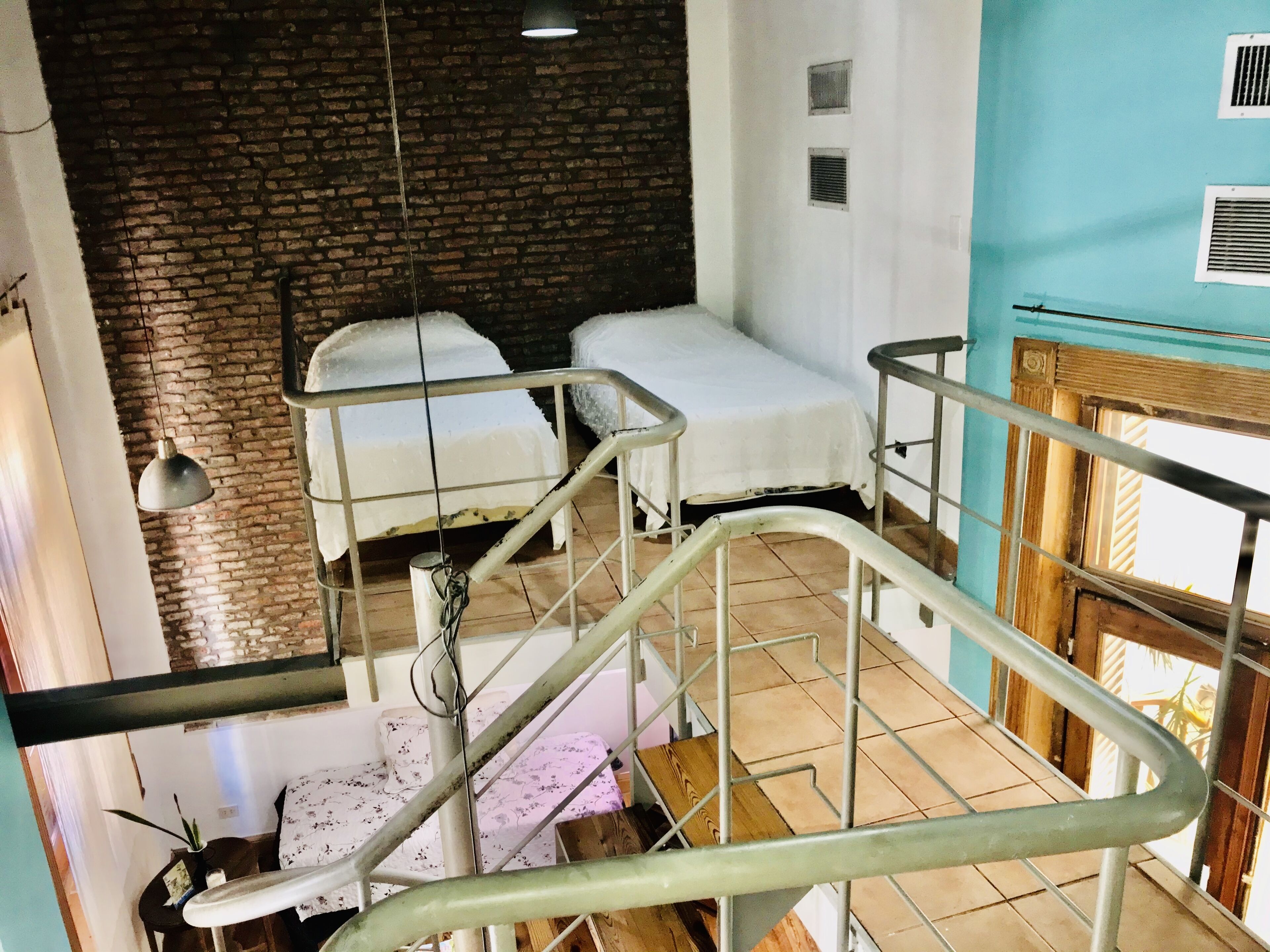 A room with two beds, a brick wall, a window with blinds, and a staircase.