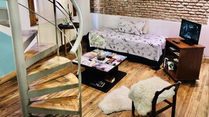 Ein modernes Wohnzimmer mit Wendeltreppe, einem Bett mit floralem Bettbezug, einem Fernseher auf einem Holzständer und einer Ziegelwand.