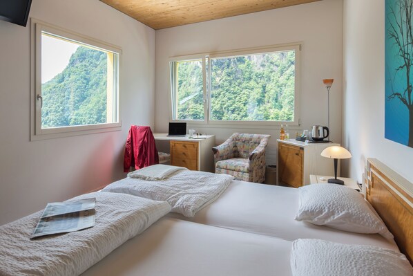 Une chambre d’hôtel avec deux lits, un bureau, une chaise, une lampe et une vue sur une montagne boisée.