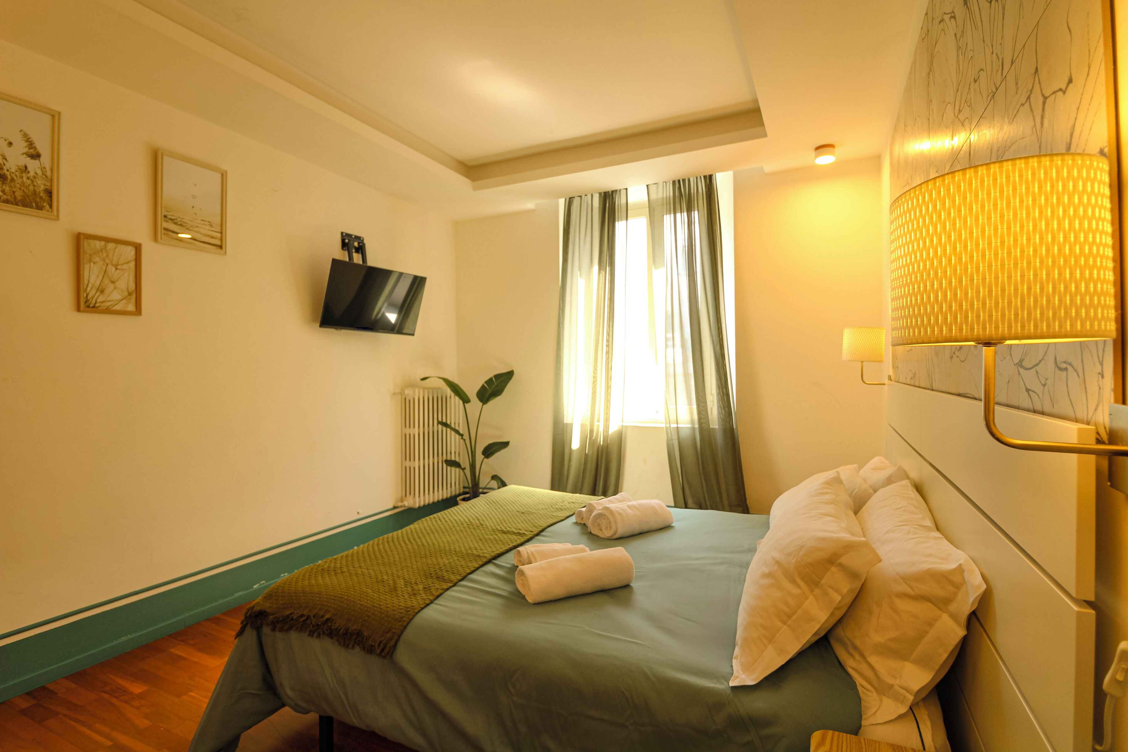 A hotel room with a bed, a television mounted on the wall, a window with curtains, and a lamp on the bedside table.
