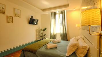 A hotel room with a bed, a television mounted on the wall, a window with curtains, and a lamp on the bedside table.
