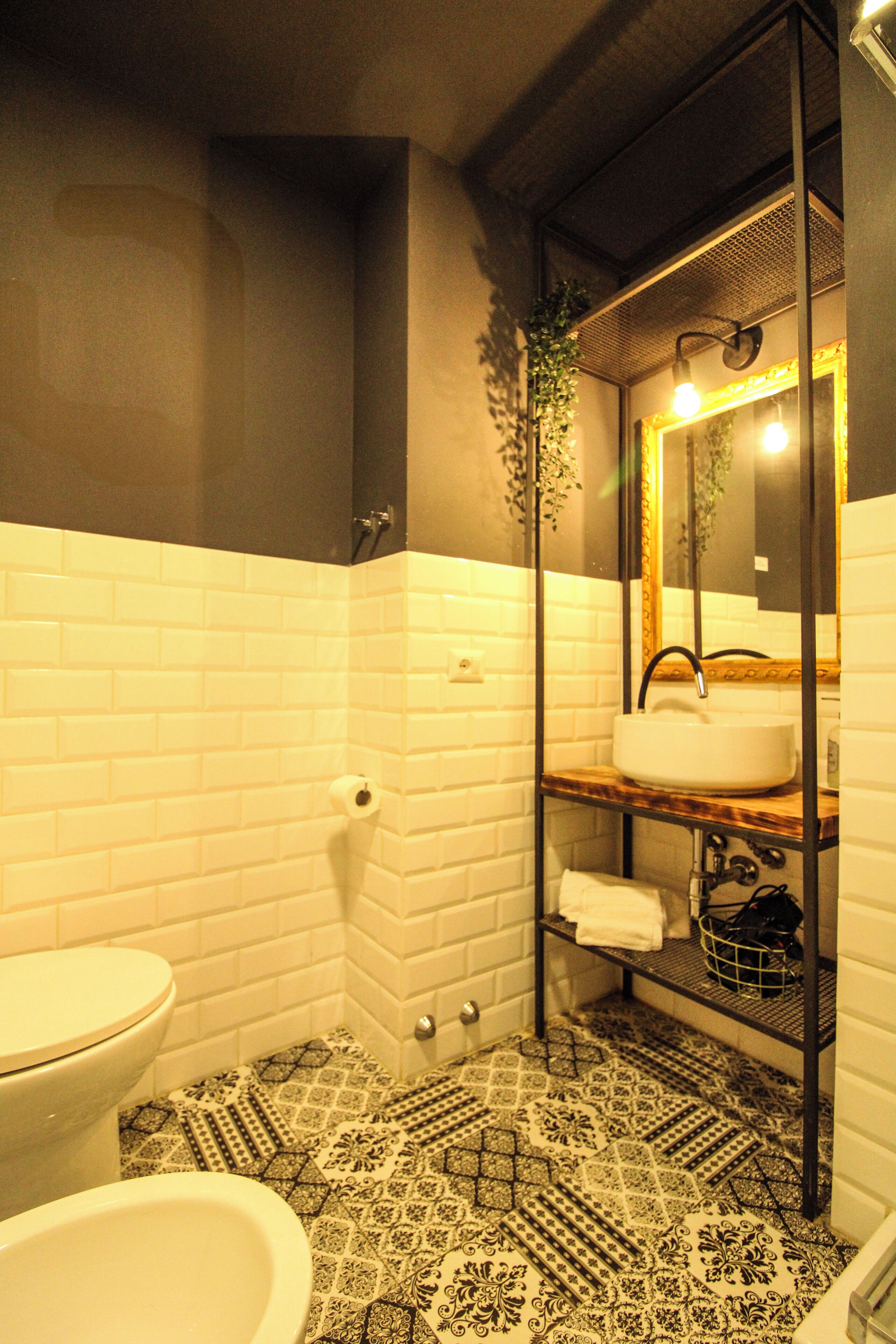 Un baño con azulejos blancos tipo metro, piso estampado, inodoro blanco, lavamanos con espejo y toallero.