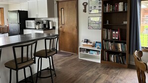 Una cocina con armarios blancos, un refrigerador negro, un microondas y una cafetera. Hay una puerta de madera, una estantería con libros y una pequeña mesa con dos sillas.