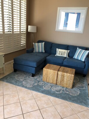 A living room with a blue sectional sofa, two striped pillows, a framed picture, and a woven ottoman. - SeaChase, 1103 East Tower, 2025 bookings now open! (Orange Beach)