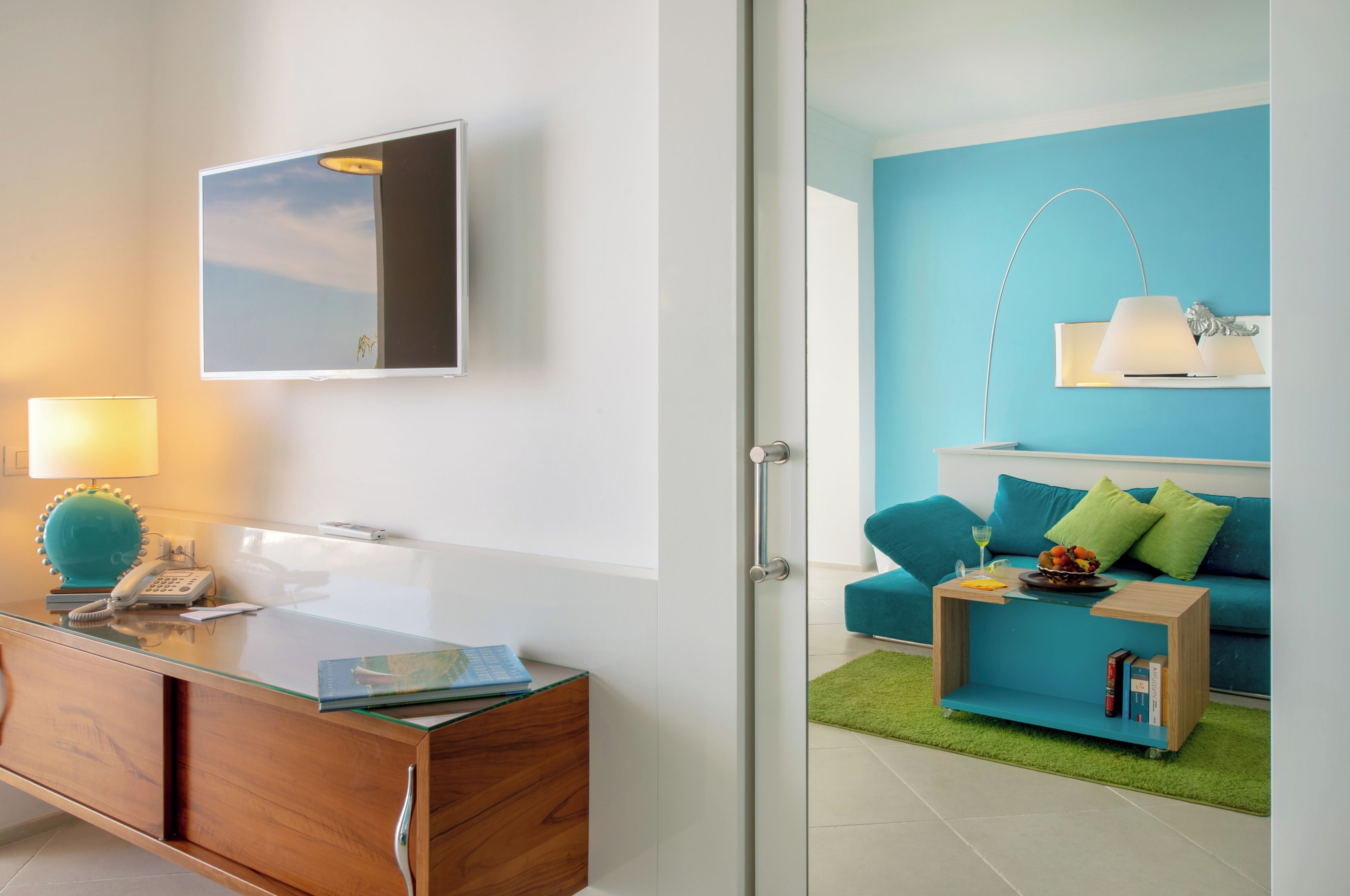 A modern room with a glass desk, a mounted TV, a blue sofa, and a small bookshelf.