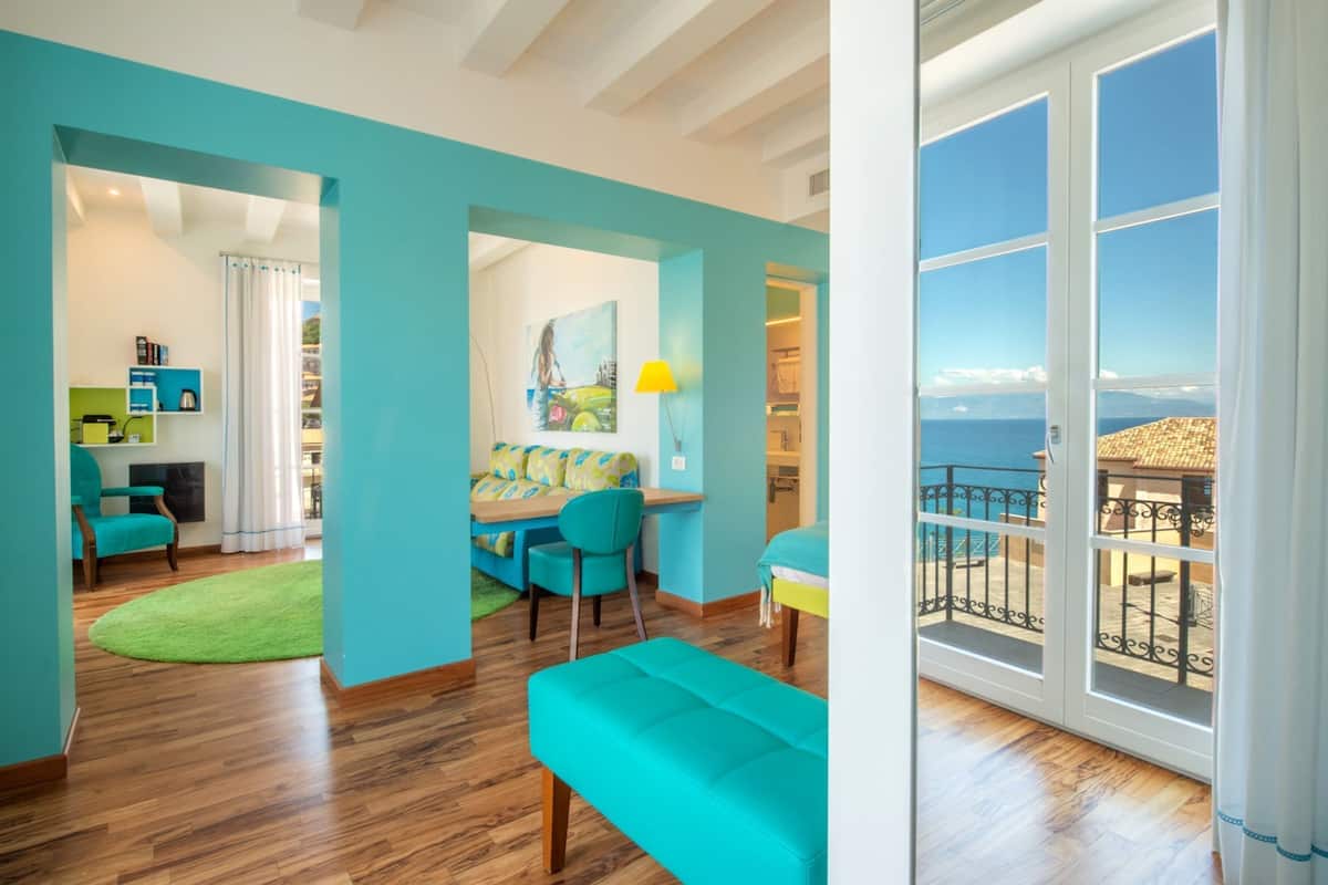A bright living room with teal walls, a wooden floor, and a sofa. There is a dining area with a table and chairs, and a balcony with a view of the sea.