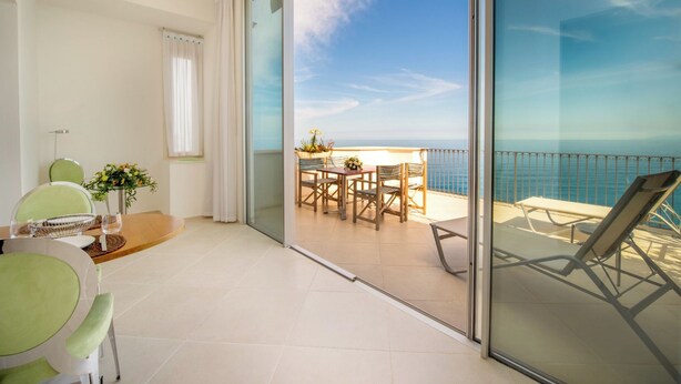 A balcony with a view of the sea, a dining area with chairs and a table, and a sliding glass door.