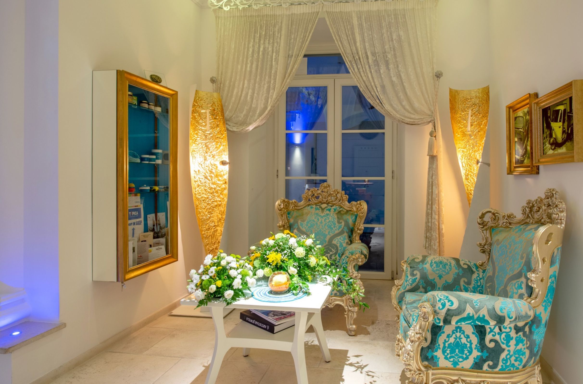 A room with a large window, ornate chairs, a table with a floral arrangement, and a wall-mounted cabinet.