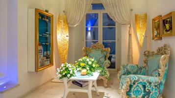 A room with a large window, ornate chairs, a table with a floral arrangement, and a wall-mounted cabinet.