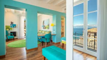 A bright room with a wooden floor, a teal sofa, a dining area with a table and chairs, and a large window with a view of the sea.