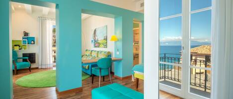 Una habitación luminosa con piso de madera, un sofá color turquesa, un comedor con mesa y sillas, y un ventanal con vista al mar.