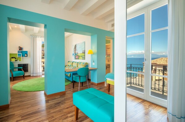A bright room with a wooden floor, a teal sofa, a dining area with a table and chairs, and a large window with a view of the sea. - Piccolo Grand Hotel (Pizzo)