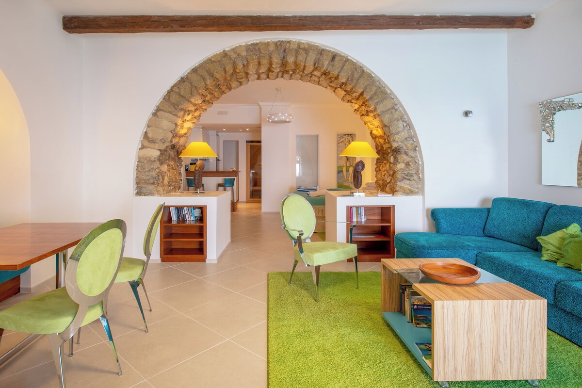 A living room with a stone archway, a blue sofa, green chairs, a wooden coffee table, and a bookshelf.