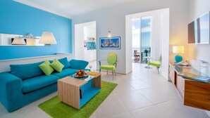 A modern living room with a blue sofa, a wooden coffee table, and a green rug. There is a dining area with a table and chairs, and a balcony with a view.