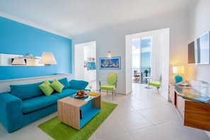 A modern living room with a blue sofa, a wooden coffee table, and a green rug. There is a dining area with a table and chairs, and a balcony with a view.