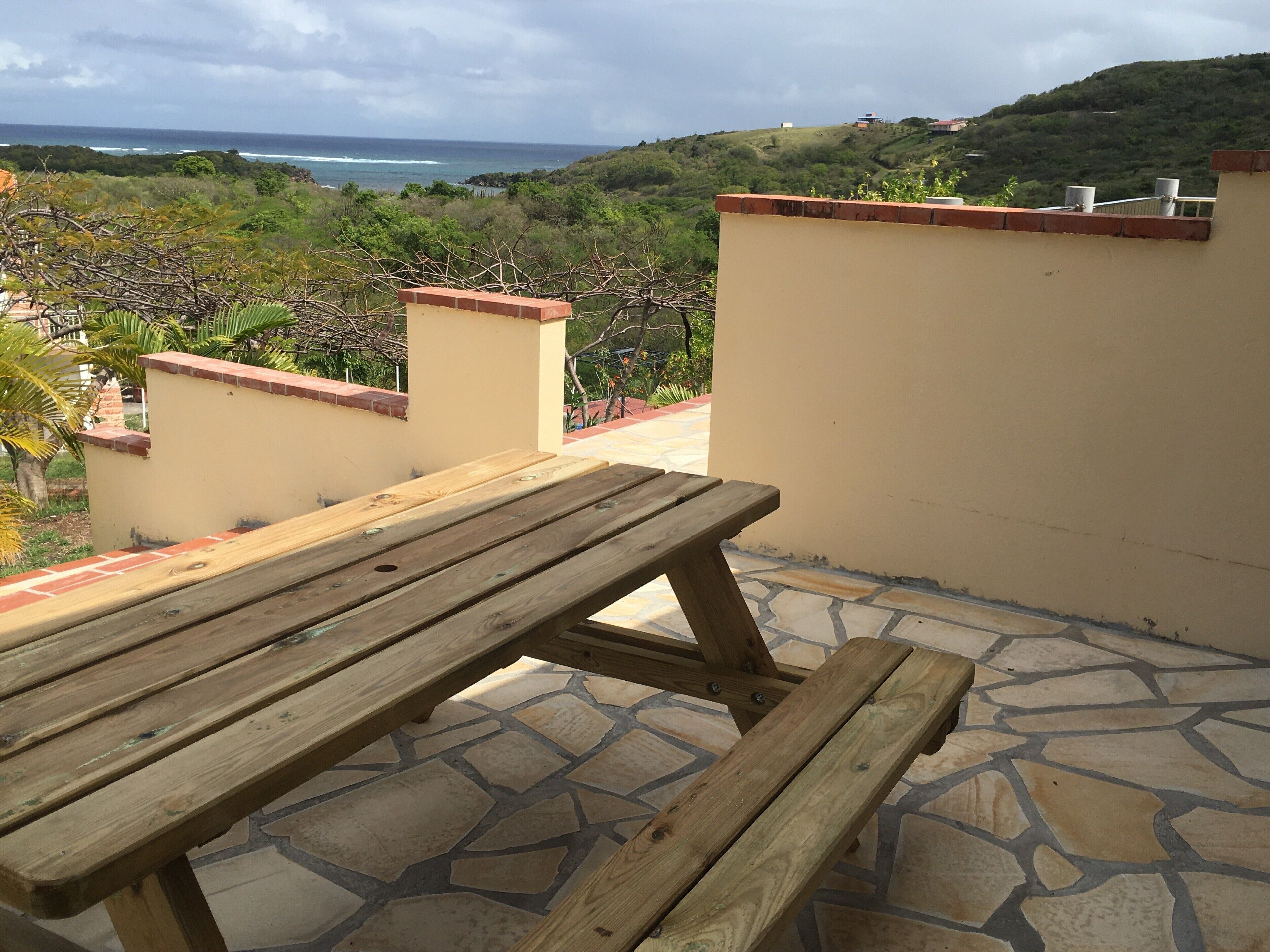 A wooden picnic table on a patio with stone flooring, overlooking a coastal landscape with hills and vegetation.