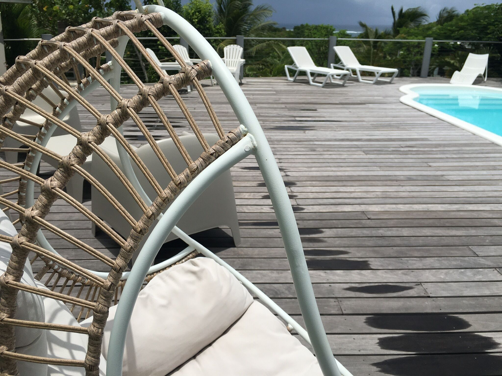 En fåtölj i rotting med en kudde, vita solstolar och en pool i bakgrunden.