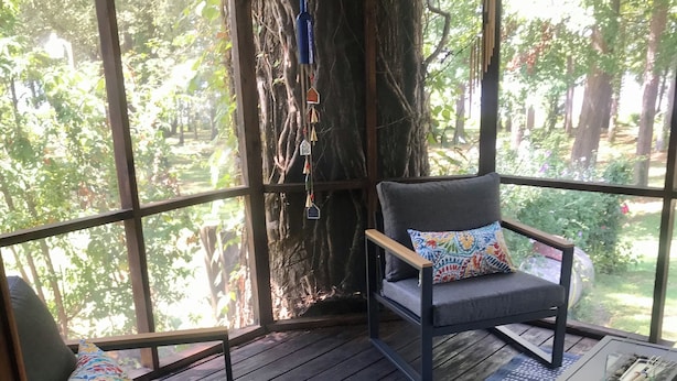 Un espacio de estar al aire libre techado con dos sillas y una mesa, rodeado por amplios ventanales y una terraza de madera.