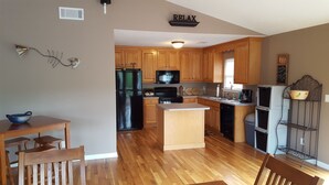 A modern kitchen with wooden cabinets, a black refrigerator, a microwave, and a stove. There is a wooden dining table and chairs, and a small island with a sink. - Lakefront Oasis: Dock, Hot Tub, Views, Nearby Rafting, Zipline, and More! (Almond)