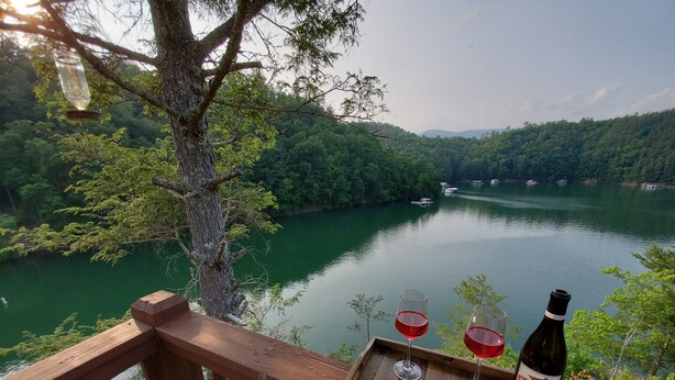A wooden deck with a tray of cheese, wine glasses, and a bottle of wine, overlooking a lake surrounded by trees. - Lakefront Oasis: Dock, Hot Tub, Views, Nearby Rafting, Zipline, and More! (Almond)