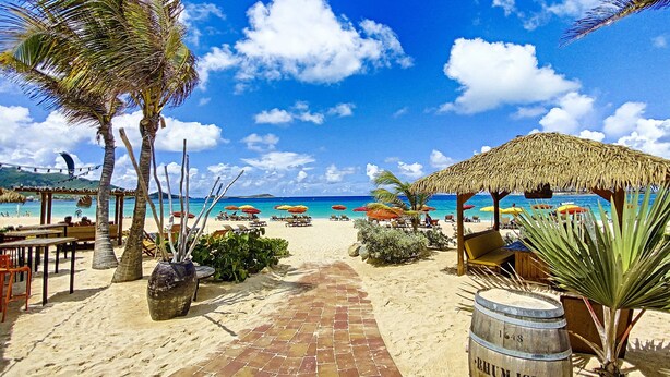 A beach scene with a paved walkway leading to the sea, palm trees, beach umbrellas, and a thatched-roof structure. - 4 Bedroom 3 Bathroom  Villa in Orient Bay, 5 min Walk to Beach and French bakery (Orient Bay)
