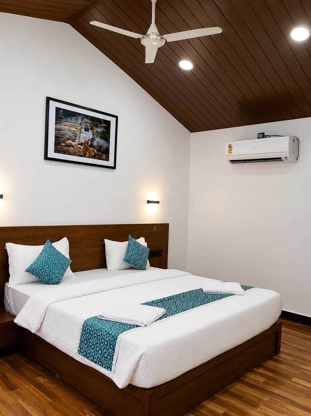 Una cama doble con cabecera de madera, ropa de cama blanca, manta estampada en color verde azulado, ventilador de techo, unidad de aire acondicionado y un cuadro en la pared.