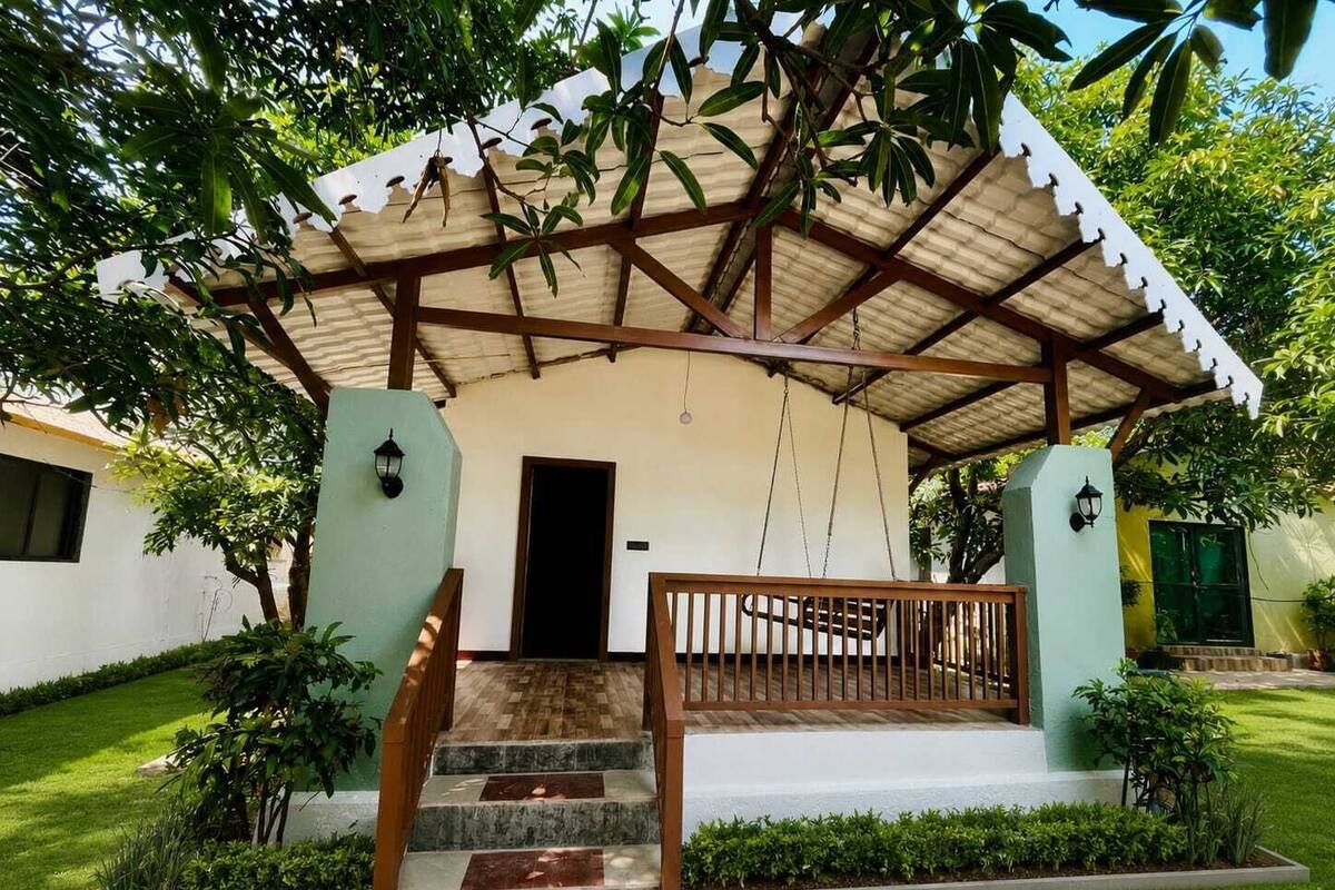 A covered outdoor area with a staircase leading to a door, surrounded by greenery and a white building.