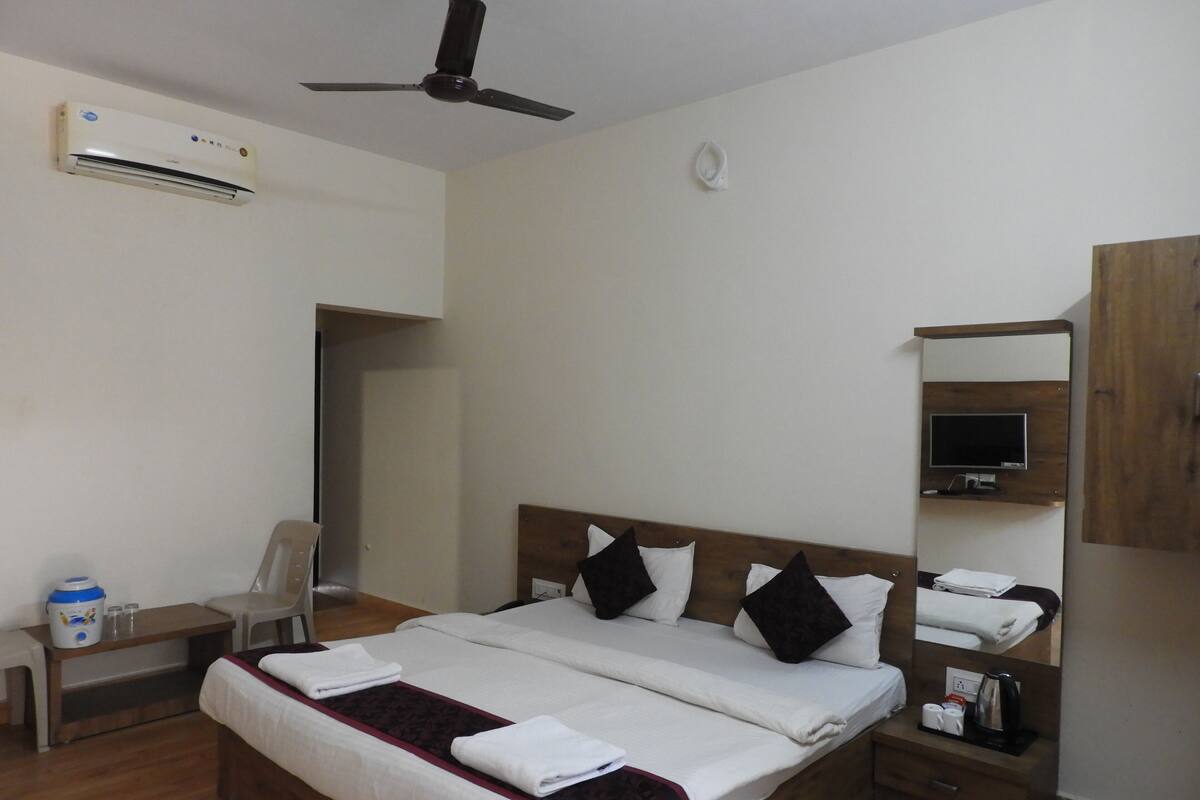 A hotel room with a bed, a desk, a chair, an air conditioner, a ceiling fan, a TV, and a mirror.
