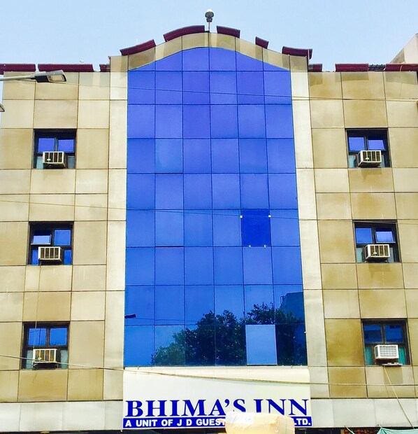 A hotel facade with a large reflective glass window, multiple air conditioning units, and a sign reading "BHIMA'S INN A UNIT OF J D GUEST TD."