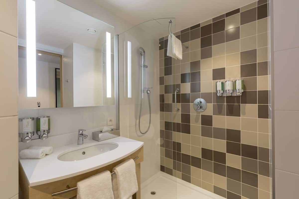 A modern bathroom with a large mirror, two sinks, a shower, and a tiled wall.