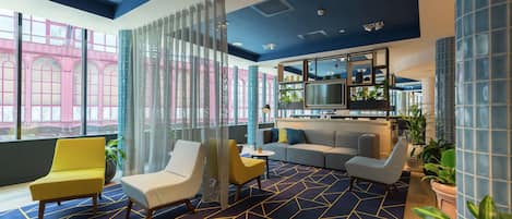 A modern lobby with a geometric patterned carpet, seating areas, a bar, and large windows with a view of a pink building.
