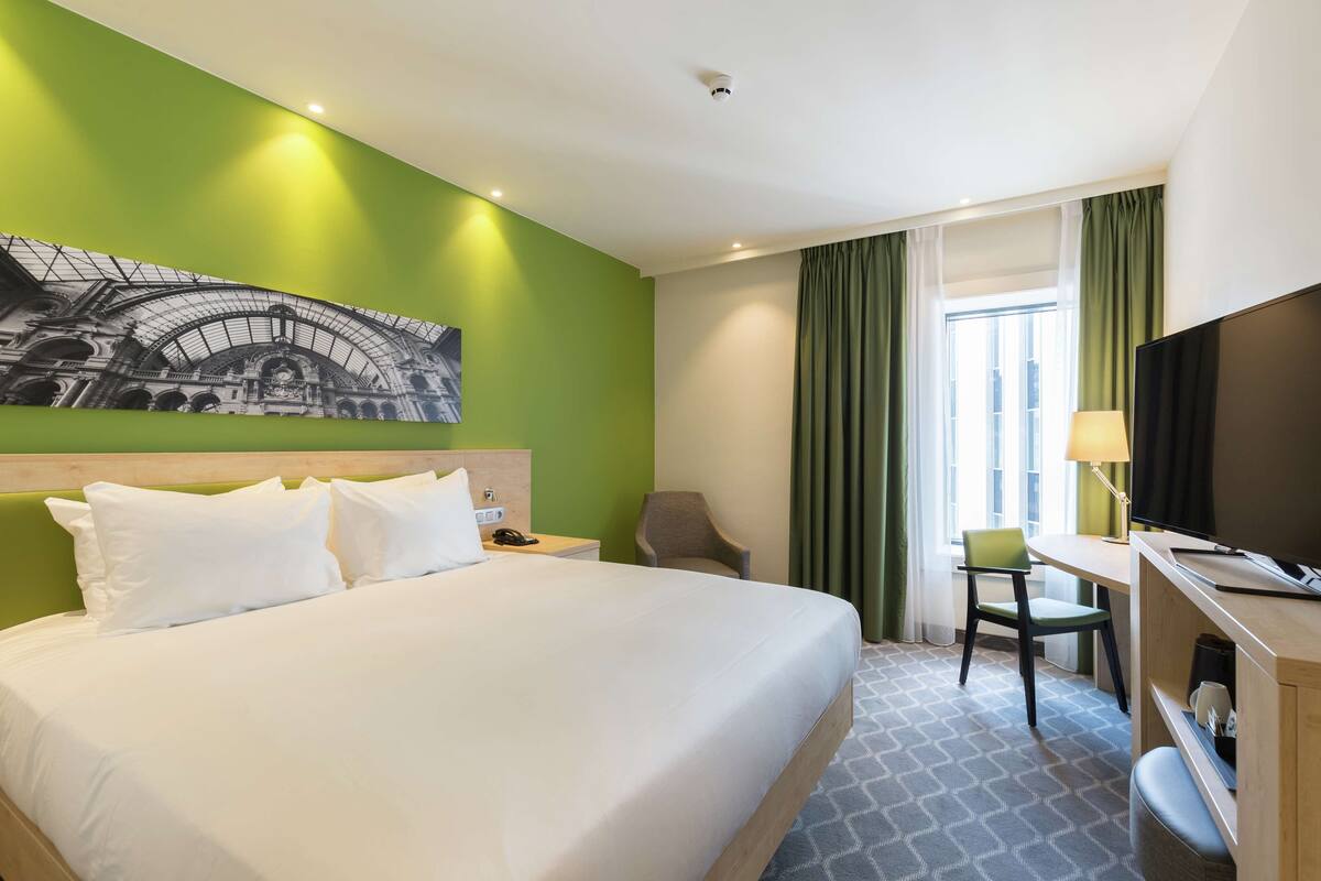 A hotel room with a large bed, a desk with a chair, a television, and a window with curtains.