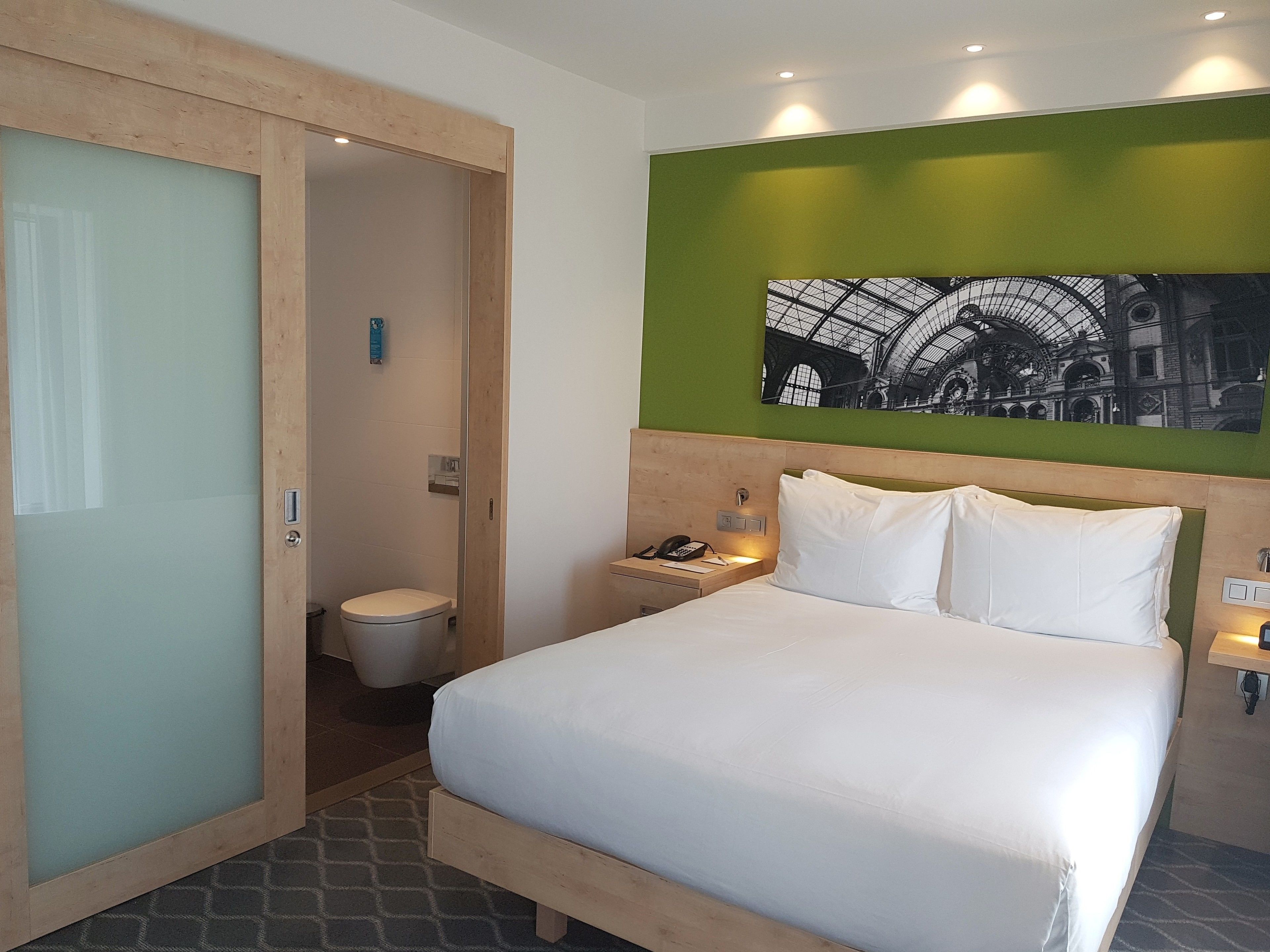 Habitación de hotel con cama, mesita de noche con teléfono, pared verde decorativa y una imagen de una estación de tren.