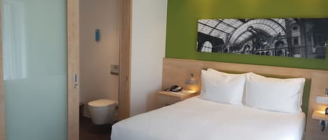 Habitación de hotel con cama, mesita de noche con teléfono, pared verde decorativa y una imagen de una estación de tren.