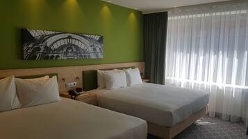 Habitación de hotel con dos camas, una pared verde, un ventanal con cortinas translúcidas y un cuadro de un edificio.