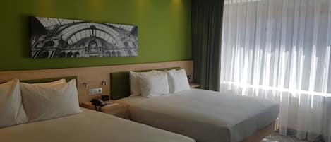 Habitación de hotel con dos camas, una pared verde, un ventanal con cortinas translúcidas y un cuadro de un edificio.
