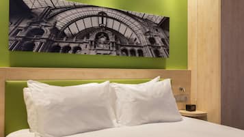 Una camera d'albergo con un letto, un poggiatesta verde, cuscini bianchi, un comodino con una radio e un quadro appeso al muro raffigurante un edificio.