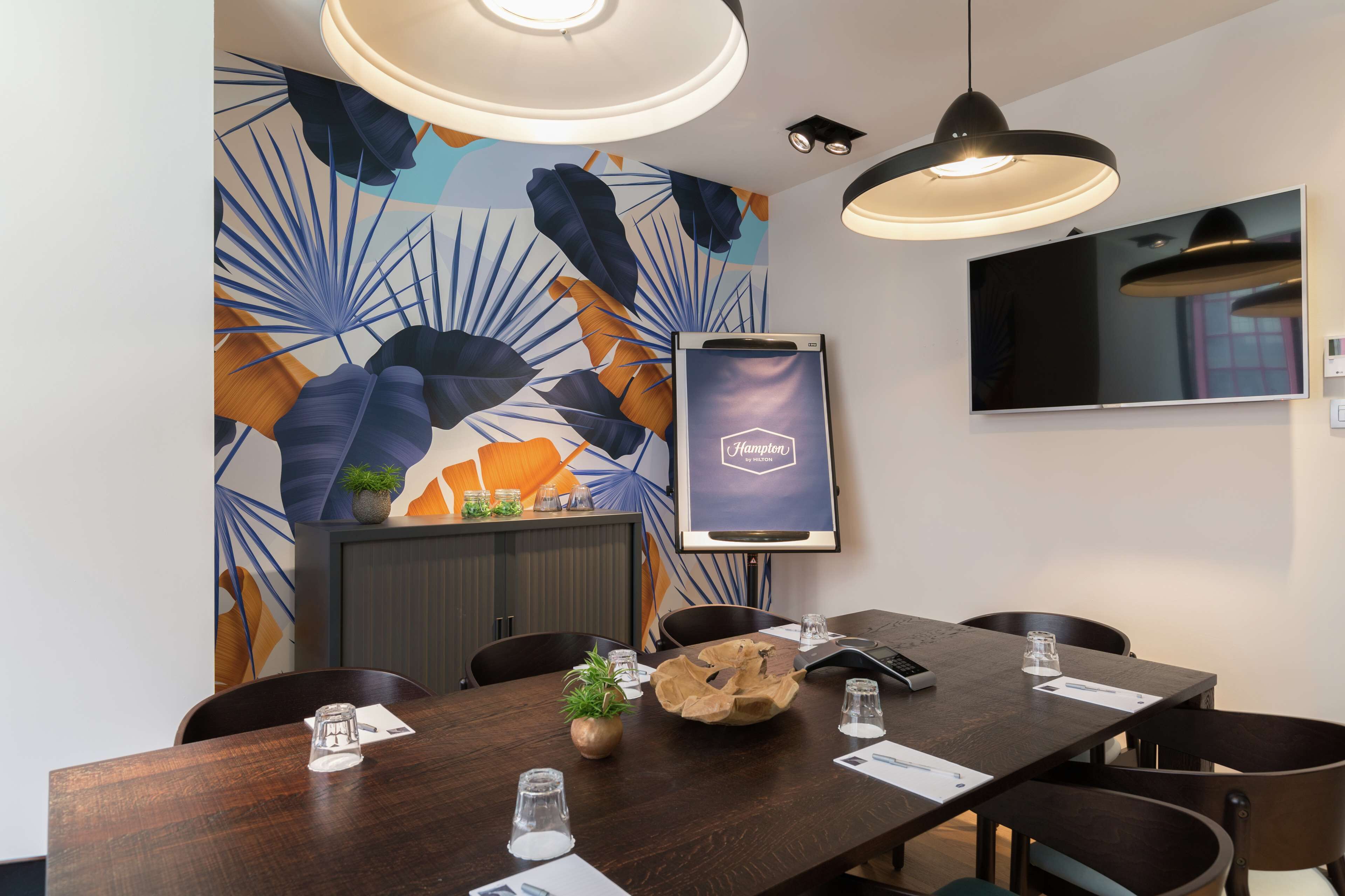 A conference room with a long table, chairs, a potted plant, and a mounted screen displaying "Hampton".