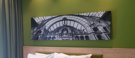 A hotel room with a bed, a nightstand with a phone, and a wall-mounted picture of a train station.