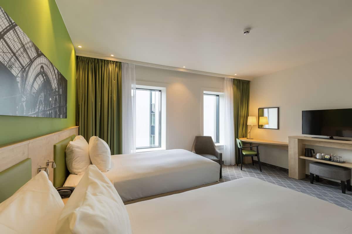 A hotel room with two beds, a desk with a chair, a television, and a large window with curtains.