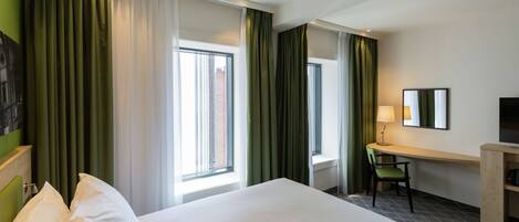 A hotel room with a bed, a desk, a chair, and two windows with curtains.