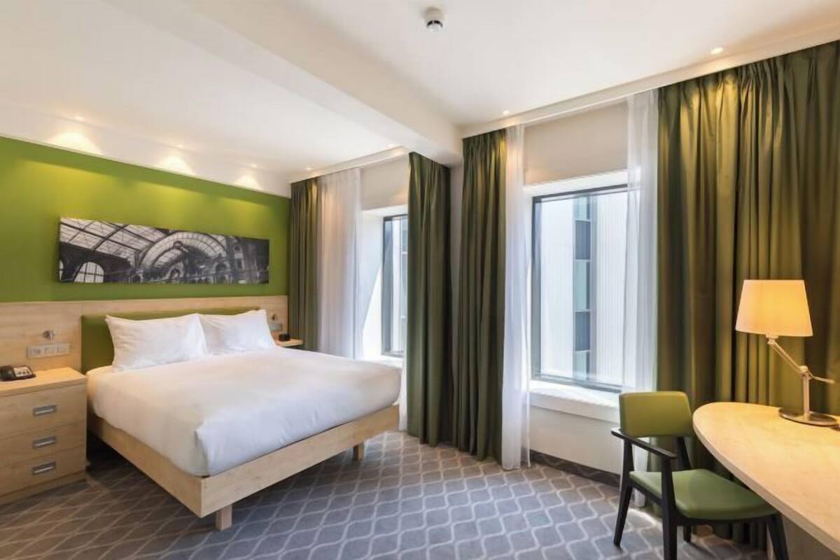 A hotel room with a bed, a desk, a chair, a lamp, a painting on the wall, and a window with curtains.